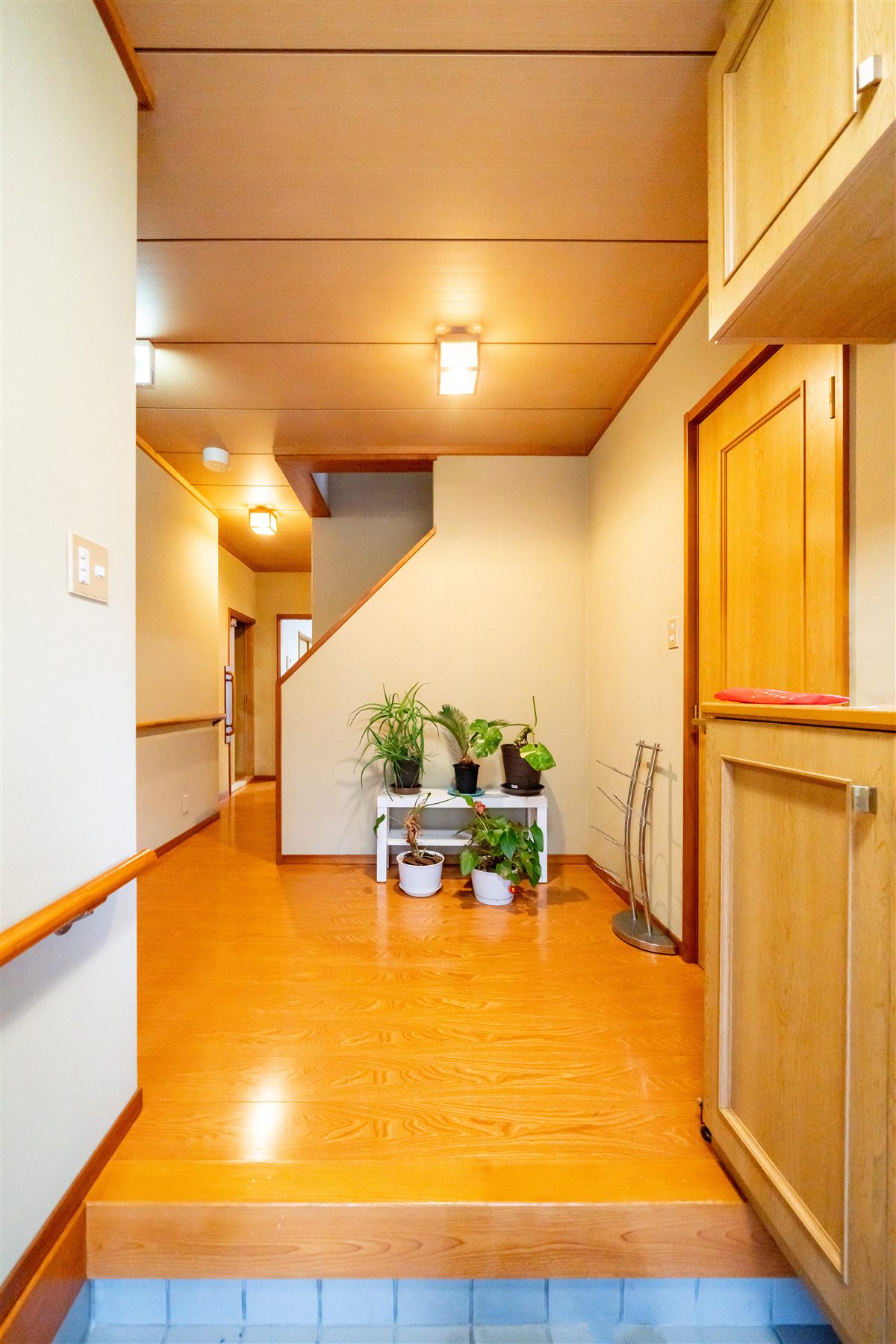 ケアホーム成田西の玄関ホール。木の床の廊下に手すりと観葉植物がある温かい空間
